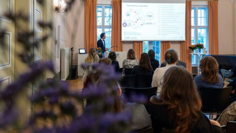 Eine männliche Person mit schwarzem Haar hält eine Präsentation am Pult (links). Rechts befindet sich das Präsentationsfeld.