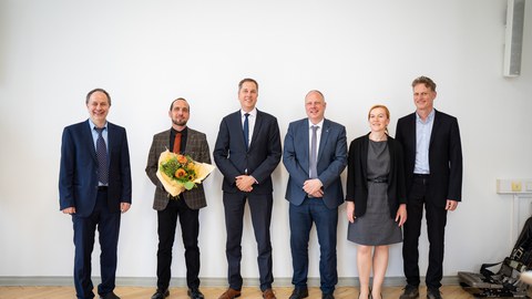 Sechs Personen stehen nebeneinander und schauen lächelnd in die Kamera. Die zweite Person von links ist Robert Kupfer. Er hält einen Blumenstrauß in der Hand.
