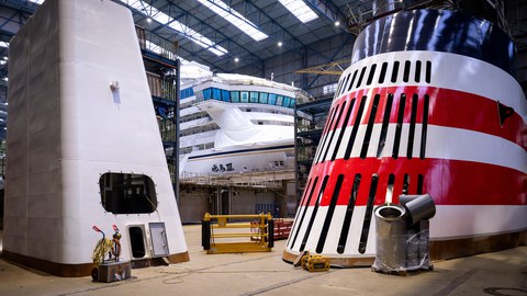 Die Hallen einer Werft. Mittig: ein großes Schiff, links und rechts weitere große Teile für den Schiffbau.
