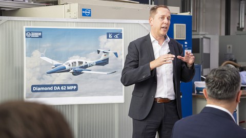Ein Mann im Anzug mit braunen Haaren spricht leidenschaftlich in einem Vortrag. Im Hintergrund befindet sich ein Plakat mit dem Forschungsflugzeug.