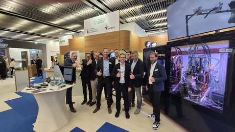 Eine Herrengruppe steht zusammen und schaut in die Kamera. Alle lachen und machen einen Daumen nach oben. Sie befinden sich auf einem Messestand. Um sie herum befinden sich Maschinen und Demonstratoren auf Tischen.