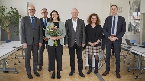 People stand in a row and look into the camera. The woman in the centre is holding a bouquet of flowers.