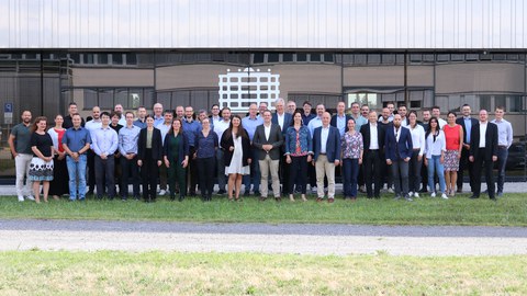 Gruppenfoto von etwa 50 Personen, die in die Kamera schauen und lächeln. Sie stehen draußen vor einem Gebäude, das im Hintergrund zu sehen ist.