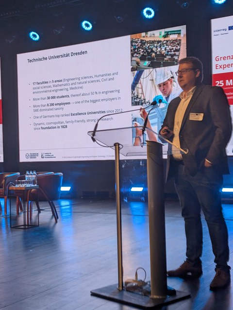 A person stands at a podium and gives a lecture. In the background, facts about TU Dresden are projected onto a huge screen.