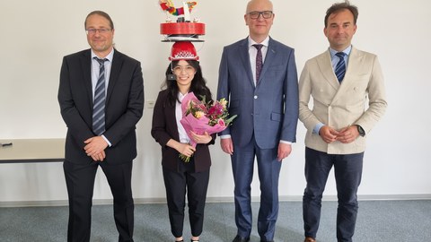 Vier Personen stehen nebeneinander, schauen in die Kamera und lächeln. Die Promovendin steht an zweiter Stelle von links und hat einen Doktorhut auf.