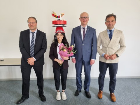 Vier Personen stehen nebeneinander, schauen in die Kamera und lächeln. Die Promovendin steht an zweiter Stelle von links und hat einen Doktorhut auf.