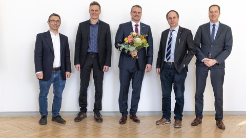 Fünf Männer im Anzug stehen in einer Reihe und lächeln in die Kamera. Die Person in der Mitte hält einen Blumenstrauß in der Hand.