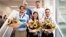 Mit Blumensträußen in der Hand blicken drei Menschen lächelnd aus der ersten Reihe in die Kamera. Dahinter stehen zwei Männer, die ebenfalls in die Kamera lächeln.