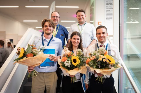 Mit Blumensträußen in der Hand blicken drei Menschen lächelnd aus der ersten Reihe in die Kamera. Dahinter stehen zwei Männer, die ebenfalls in die Kamera lächeln.