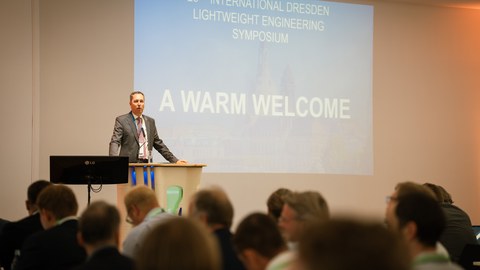 A man stands at a lectern and speaks. Many people are sitting in the foreground listening to him. You can see them from behind.