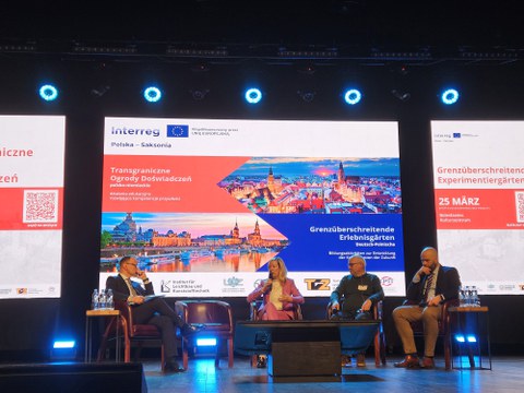 Four people are sitting on a stage in a semicircle. A woman speaks into a microphone. In the background is a large screen with pictures of the cities of Dresden and a Polish city.