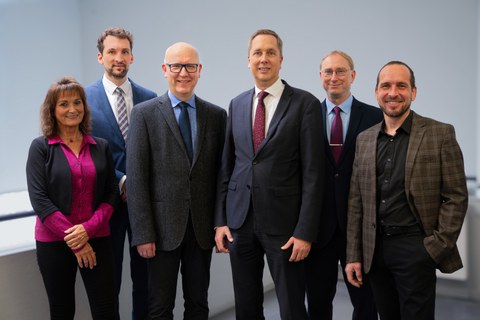 Portrait von sechs Personen: eine Frau in einem pinken Hemd links außen lächelt in die Kamera, es folgen sechs Männer im Anzug, welche ebenfalls freundlich in die Kamera lächeln.