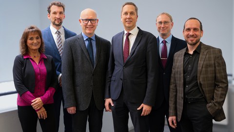Portrait of six people: a woman in a pink shirt on the far left smiles at the camera, followed by six men in suits, who also smile warmly at the camera.