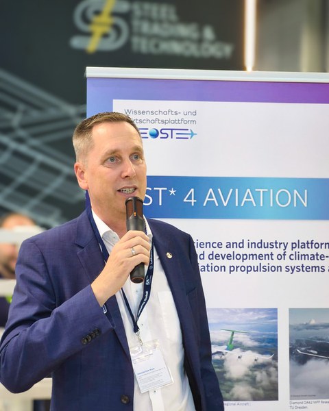 Prof. Gude, a man in a suit, is speaking into a microphone. Behind him is an OST4Aviation roll-up banner.