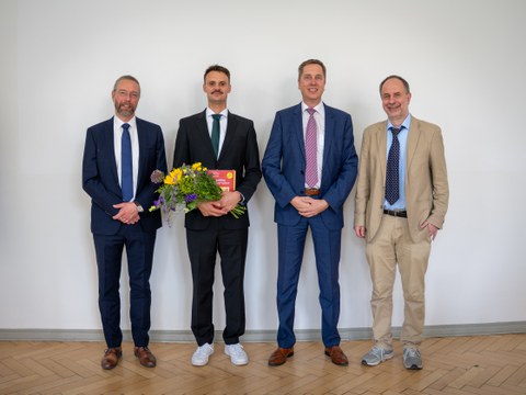 Four men in suits stand side by side, smiling at the camera. The second man from the left is holding a bouquet of flowers.