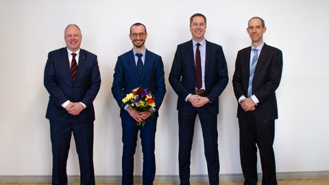 Vier Männer in Anzügen stehen in einer Reihe und lächeln in die Kamera. Die zweite Person von links hält einen Blumenstrauß in der Hand.