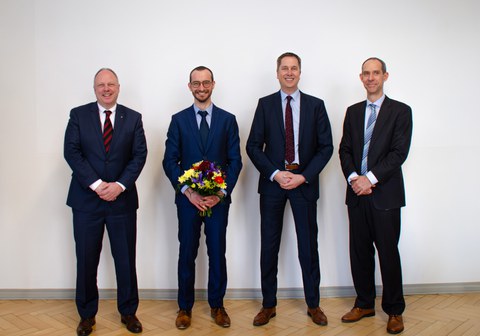 Vier Männer in Anzügen stehen in einer Reihe und lächeln in die Kamera. Die zweite Person von links hält einen Blumenstrauß in der Hand.
