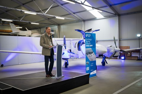 A man with gray hair and a coat is standing on a stage in front of a lectern, speaking into a microphone. In the background is an airplane, festively lit, and a roll-up banner.
