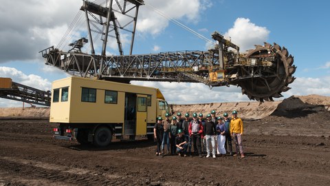 A group of people stands in the open-cast mine area. A mining vehicle can be seen in the background.