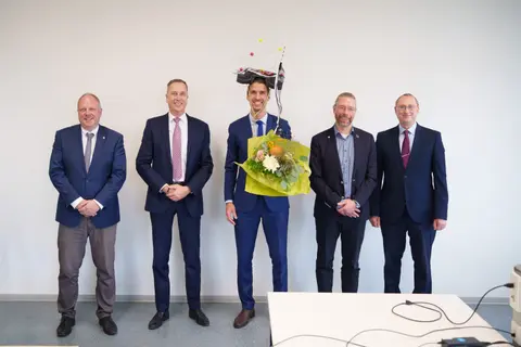 Fünf Männer im Anzug stehen in einer Reihe und lachen in die Kamera. In der Mitte befindet sich Dr.-Ing. Daniel Köhler mit seinem Promotionshut sowie einem Blumenstrauß.