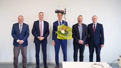 Five men in suits are standing in a row, smiling at the camera. In the middle is Dr.-Ing. Daniel Köhler, wearing his doctoral cap and holding a bouquet of flowers.