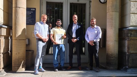 Andreas Hornig, Dr. Rohan Soman, Prof. Dr. Maik Gude und Dipl.-Ing. Philip Steinbild bei der Überreichung der Urkunde: Vier Personen stehen vor einem Gebäude auf einer Treppe und lächeln in die Kamera. Dr. Soman hält eine Urkunde in der Hand.