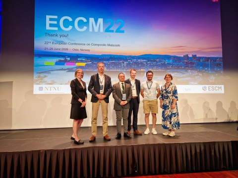 Auf einer Bühne stehen sechs Personen, die in die Kamera schauen. Hinter ihnen wird eine ECCM-22-Folie projiziert.