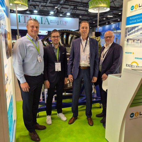 Four men wearing shirts and suits stand side by side on the exhibition floor, smiling at the camera.