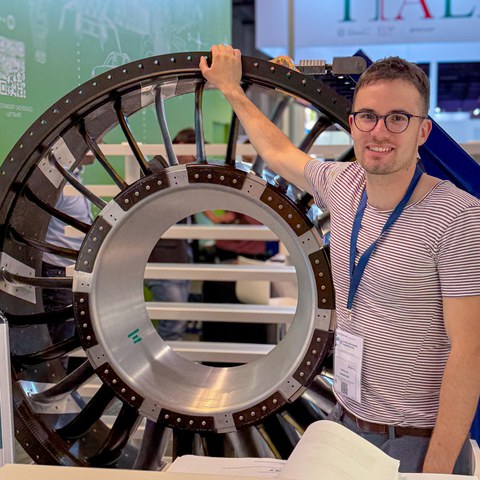 A smiling man wearing glasses stands at an exhibition booth next to a circular aircraft engine component. The exhibit is taller than the person.