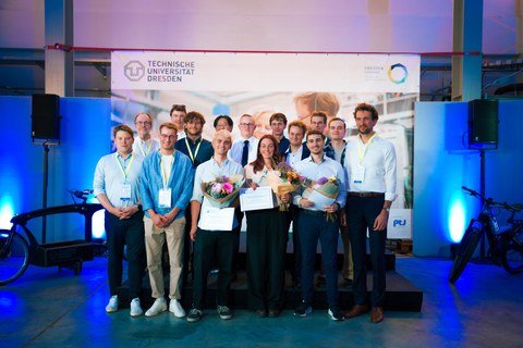 Auf einer Bühne steht eine Gruppe von Student:innen beieinander und lächelt in die Kamera. Die Gewinner:innen halten Blumensträuße in den Händen