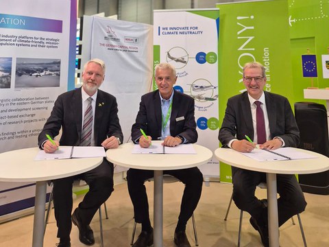 Three men in suits are sitting at a table and signing the MoC.
