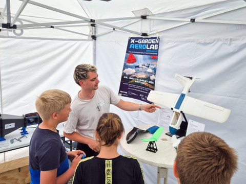 Ein junger Ingenieur steht im Pavillon und zeigt auf ein Modellflugzeug, während Kinder um ihn herum stehen und zuhören.