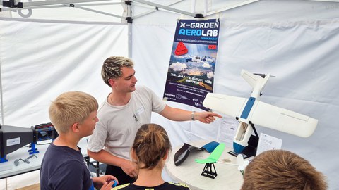 Ein junger Ingenieur steht im Pavillon und zeigt auf ein Modellflugzeug, während Kinder um ihn herum stehen und zuhören.