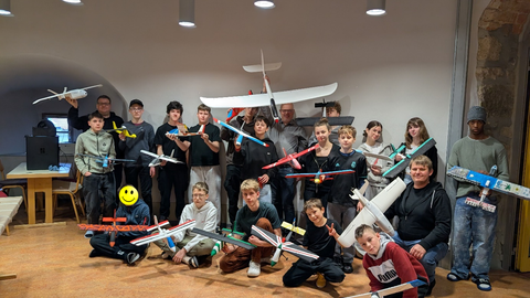 Gruppenbild von Schüler:innen und betreuenden Ingenieur:innen beim X-Garden AeroLab. Die Teilnehmenden lächeln in die Kamera und präsentieren ihre selbst konstruierten und gebauten Modellflugzeuge.
