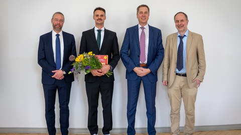 Four men in suits stand side by side, smiling at the camera. The second man from the left is holding a bouquet of flowers.