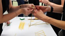 Student:innen bauen aus Spaghetti einen Turm. Hände halten die Spaghetti in der Hand, welche teilweise mit Klebeband miteinander verbunden sind.