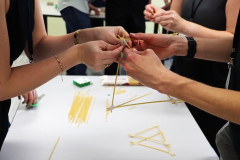 Student:innen bauen aus Spaghetti einen Turm. Hände halten die Spaghetti in der Hand, welche teilweise mit Klebeband miteinander verbunden sind.
