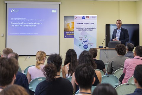 Ein Mann steht vor einem Pult und spricht vor Menschen, die im Publikum sitzen. Im Hintergrund ist eine Folie an die Wand proijziert.