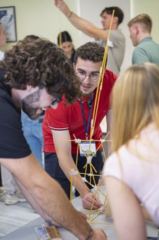 Im Vordergrund ist ein Spaghettiturm zu sehen, der durch Klebeband verbunden ist. Im Hintergrund ist ein Student zu sehen, der sich über den Tisch lehnt.