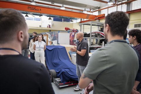 Student:innen stehen im Halbkreis und schauen einen glatzköpfigen Mann an, welcher etwas erzählt. Sie befinden sich in einer Produktionshalle o.ä.