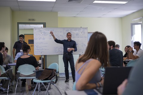 Ein Mann steht vor einer Tafel und erklärt den Student:innen, die drum herum sitzen, gestikulierend etwas.