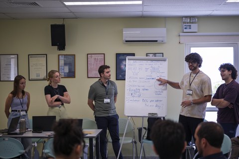 Student:innen stehen vor einer beschriebenen Tafel. Einer von ihnen erklärt dem Publikum gestikulierend den Inhalt der Tafel.