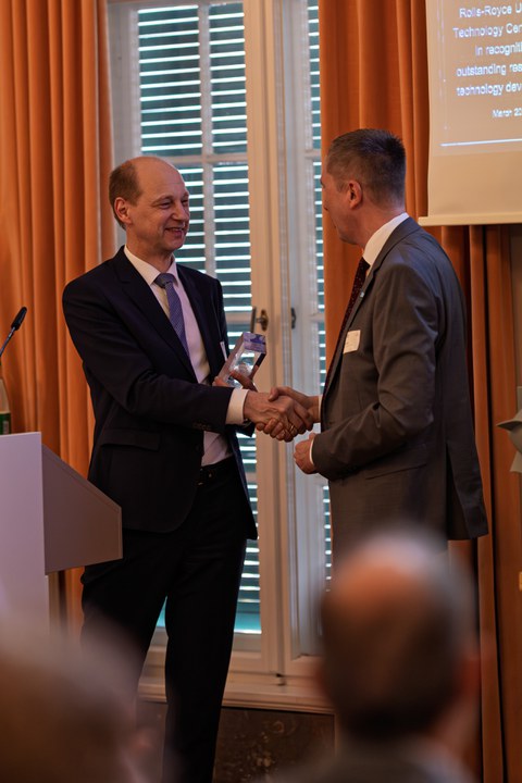 Two men in suits are standing facing each other. They shake hands with their right hands. The man on the left (Mr. Roland Wilhelm) is holding a trophy, which he wants to present to the other man (Prof. Gude). 