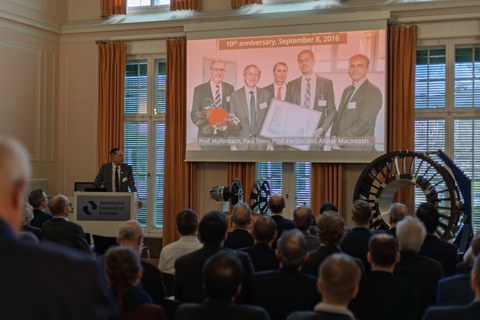 A man is standing at a lectern; a group photo of four men is projected onto a screen. Above it is the headline “10th anniversary, September 8, 2016.” The audience is watching the scene in front of them. Demonstrators are standing in the right-hand corner.