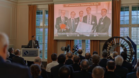 A man is standing at a lectern; a group photo of four men is projected onto a screen. Above it is the headline “10th anniversary, September 8, 2016.” The audience is watching the scene in front of them. Demonstrators are standing in the right-hand corner.