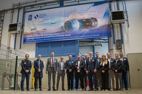 People in business attire are lined up under a banner in a technical setting. The banner features a futuristic aircraft and the caption “More than 20 years of trusted partnership and outstanding research and technology...”.