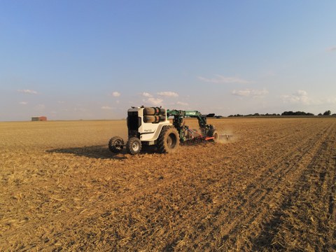 Auf dem Bild ist ein autonomer Feldroboter bei der Bodenbearbeitung auf einem großen, abgeernteten Acker zu sehen. Das vierrädrige Fahrzeug fährt mittig durch das Feld und zieht ein Bodenbearbeitungsgerät hinter sich her, das eine leichte Staubwolke erzeu