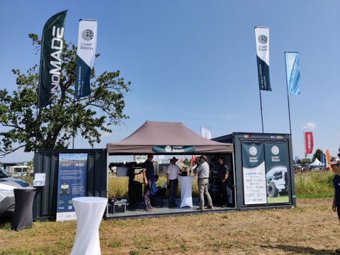 Das Bild zeigt einen Messestand auf einem Freigelände, vermutlich auf einer Messe für Landtechnik. Der Stand besteht aus einem umgebauten Container und einem Zelt, unter dem sich mehrere Personen befinden. Auf dem Container und dem Zelt sind diverse Werbebanner angebracht,
