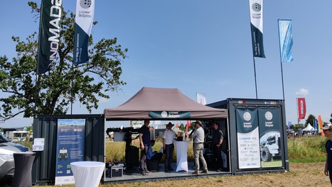 Das Bild zeigt einen Messestand auf einem Freigelände, vermutlich auf einer Messe für Landtechnik. Der Stand besteht aus einem umgebauten Container und einem Zelt, unter dem sich mehrere Personen befinden. Auf dem Container und dem Zelt sind diverse Werbebanner angebracht,