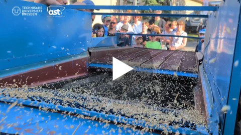 Besucherinnen und Besucher schauen gespannt auf eine landwirtschaftliche Maschine, die Getreide aussiebt, während Körner durch die Luft fliegen. Aufnahme von der Langen Nacht der Wissenschaft an der TU Dresden.